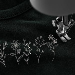 May include: A close-up of a black fabric with embroidered white floral designs. A sewing machine needle is positioned above the fabric, suggesting the creation of custom apparel or home decor items.