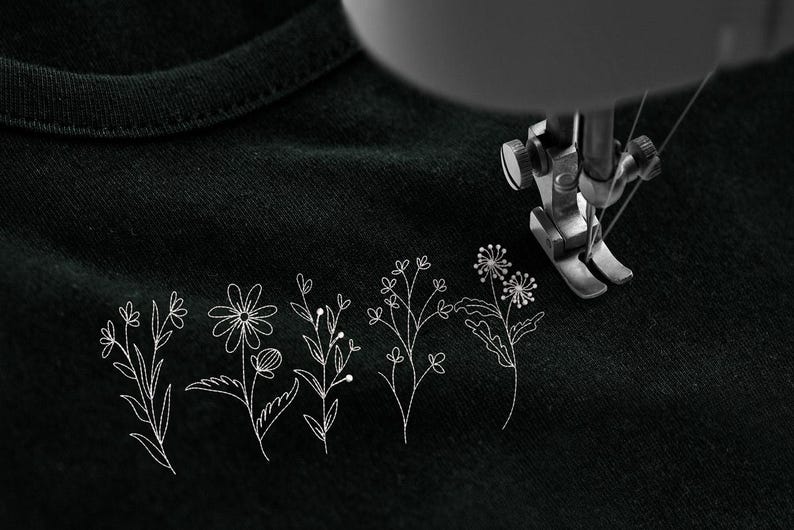 May include: A close-up of a black fabric with embroidered white floral designs. A sewing machine needle is positioned above the fabric, suggesting the creation of custom apparel or home decor items.