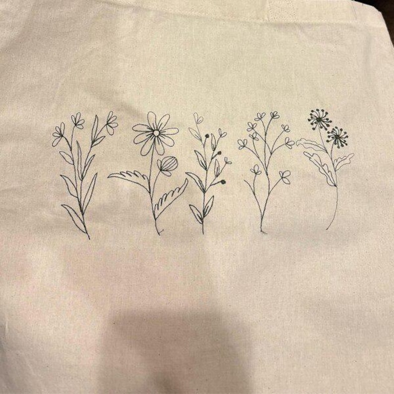 May include: Cream-colored tote bag featuring a black line drawing of five different floral stems. The flowers are in various stages of bloom, with detailed leaves and petals. The bag is made of a sturdy fabric.