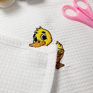 May include: A white waffle-weave fabric with an embroidered yellow duckling peeking out. The duckling has a yellow body, orange beak, and black eyes. Pink scissors are in the background.