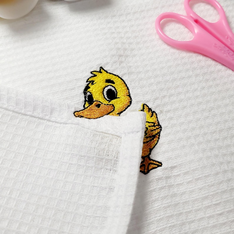 May include: A white waffle-weave fabric with an embroidered yellow duckling peeking out. The duckling has a yellow body, orange beak, and black eyes. Pink scissors are in the background.