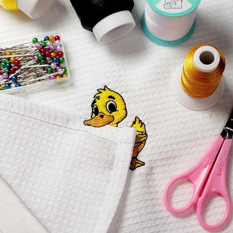 May include: White waffle-weave towel with an embroidered yellow duckling design. Sewing supplies include spools of black, white, and gold thread, colorful pins, and pink scissors. A craft project in progress.