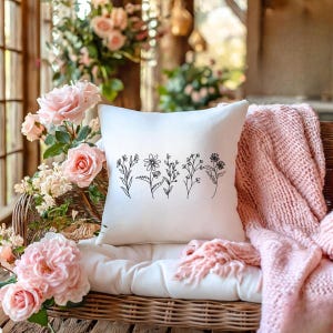 May include: A square white pillow with a black line drawing of various wildflowers. The pillow is on a wicker chair with a white cushion and a pink knitted throw. Pink roses and other flowers are in the background.