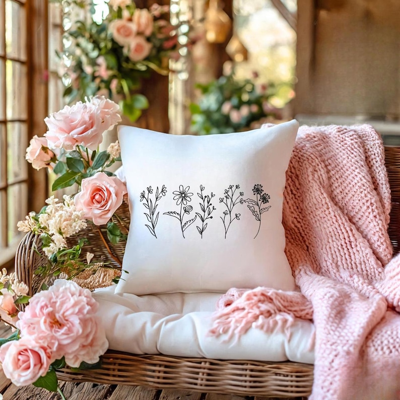 May include: A square white pillow with a black line drawing of various wildflowers. The pillow is on a wicker chair with a white cushion and a pink knitted throw. Pink roses and other flowers are in the background.