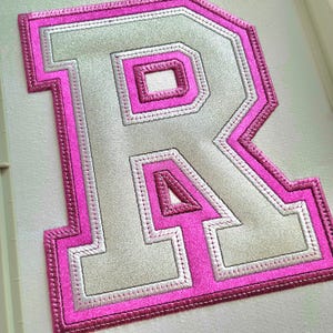 May include: Embroidered letter "R" patch in a varsity style. The letter is light gray with a pink border and accents. The patch is on a white background.