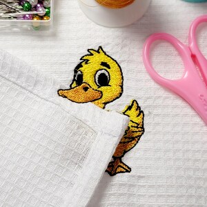 May include: Embroidered yellow duckling peeking out from a white waffle-weave fabric. The duckling has a yellow body, orange beak, and black eyes. Pink scissors and sewing supplies are in the background.