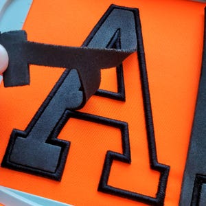 Könnte beinhalten: Nahaufnahme eines schwarzen Buchstaben "A" Applikation mit schwarzer gestickter Umrandung, teilweise von einem orangefarbenen Stoff abgezogen. Die Applikation besteht aus einem weichen, filzartigen Material.