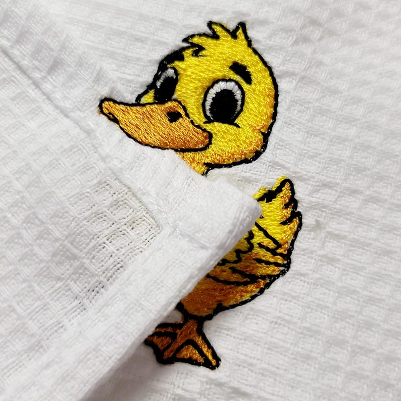 May include: White waffle-weave fabric with an embroidered yellow duckling design. The duckling has a yellow body, orange beak and feet, and black eyes. The fabric is folded over, partially obscuring the duckling.