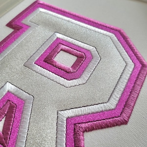 以下が含まれることがあります： 銀色の中心とピンクと白の刺繍の縁取りが付いた文字「P」のパッチのクローズアップ。パッチはテクスチャ加工された外観で、白い背景に配置されています。デザインは、レイヤー化された幾何学的なスタイルが特徴です。