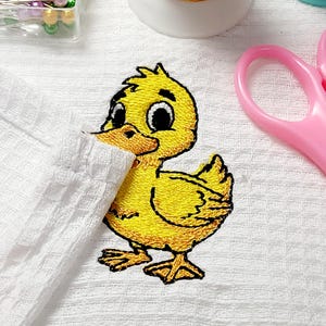 May include: A yellow embroidered duckling patch with black outlines and large eyes. The duckling is on a white textured surface, with a pair of pink scissors and sewing supplies in the background. The patch is a craft supply.
