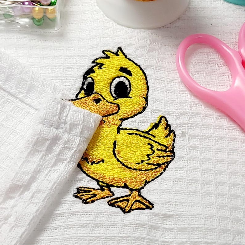 May include: A yellow embroidered duckling patch with black outlines and large eyes. The duckling is on a white textured surface, with a pair of pink scissors and sewing supplies in the background. The patch is a craft supply.
