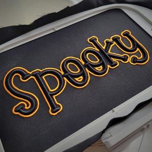 May include: Black fabric with the word "Spooky" embroidered in a stylized font. The word is outlined in black with a bright orange fill, creating a bold, eye-catching design. The embroidery is set within a white frame.