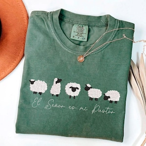 Op de afbeelding: Groen Comfort Colors T-shirt met geborduurd schapenontwerp en de tekst "El Señor es mi Pastor". Een gouden ketting met een hanger ligt op het shirt. Een bruine hoed en een gedroogde plant staan op de achtergrond.