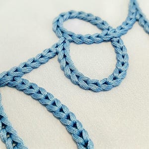 May include: A light blue braided cord is arranged in a cursive script pattern on a smooth, light-coloured fabric surface. The cord features a distinct, repeating chain-link texture created by the intricate weaving of the blue thread.
