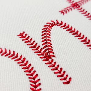 May include: Close-up of white fabric with red embroidered stitching forming the word "HOME". The stitching is in a baseball-like pattern, with the red thread creating a bold contrast against the white background. The image suggests a sports-themed or decorative item.