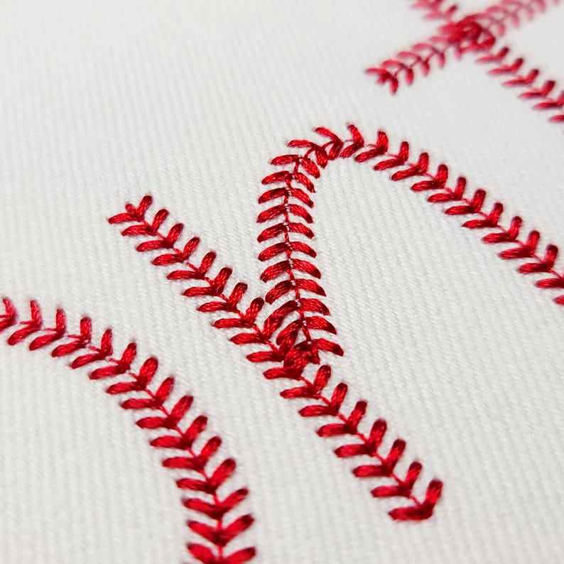 May include: Close-up of white fabric with red embroidered stitching forming the word "HOME". The stitching is in a baseball-like pattern, with the red thread creating a bold contrast against the white background. The image suggests a sports-themed or decorative item.