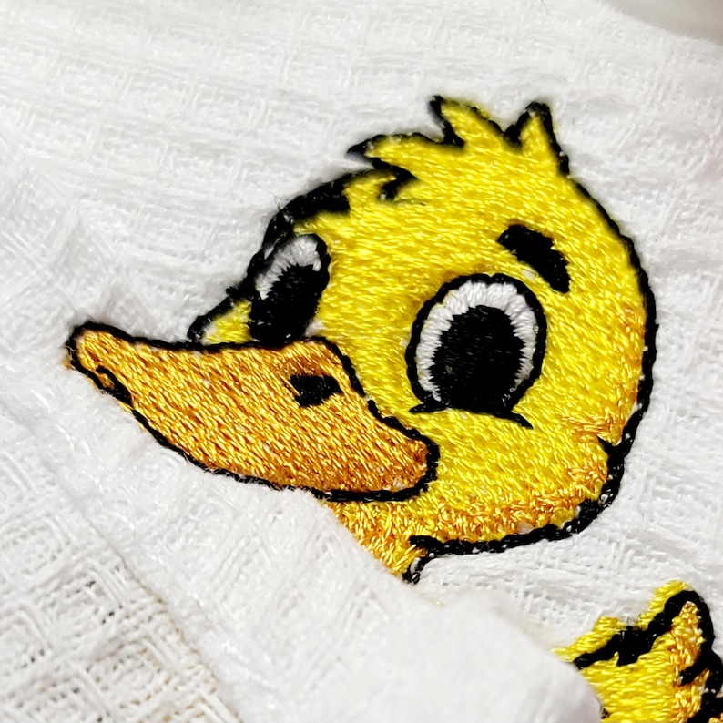 May include: Close-up of a white fabric with an embroidered yellow duckling design. The duckling has black eyes, a yellow beak, and black outlines. The embroidery is detailed, and the fabric has a textured appearance.