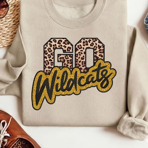 May include: A beige sweatshirt featuring the text "GO Wildcats". The word "GO" is in a leopard print, and "Wildcats" is in a gold glitter font. The sweatshirt has a crew neck and long sleeves, suitable for casual wear.