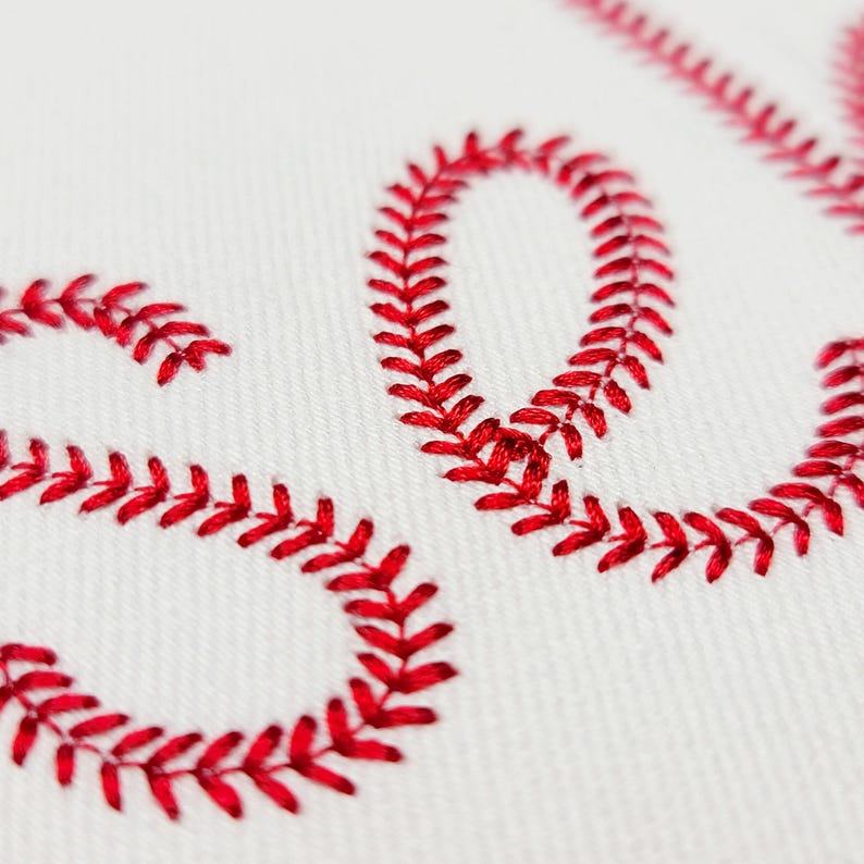 May include: Close-up of white fabric with red baseball-style stitching forming the letters "SO". The stitching is detailed and creates a textured effect against the smooth white background. The letters are part of a larger word.