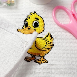 May include: Embroidered yellow duckling patch with black details and an orange beak, on a white waffle-weave fabric. The duckling is a cheerful design, perfect for adding to clothing or accessories. Pink scissors are in the background.