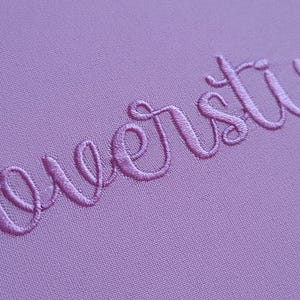 May include: Close-up of a lavender fabric with embroidered cursive text. The thread colour matches the fabric, creating a subtle, textured effect. The image highlights the detailed stitching and the soft, muted colour of the material.