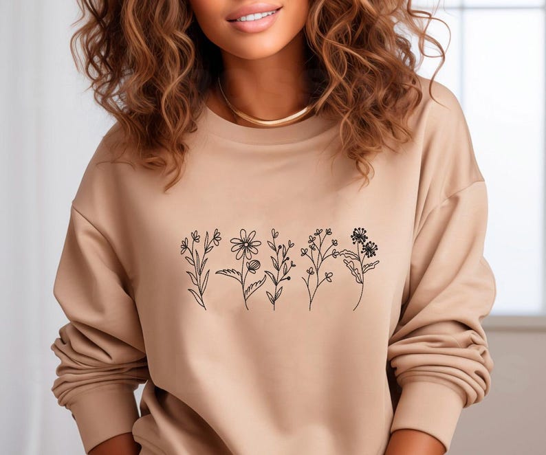 May include: A tan-colored sweatshirt featuring a minimalist floral design in black ink. The design includes various stylized flowers and stems. The sweatshirt has long sleeves and a crew neck. The overall style is casual and comfortable.