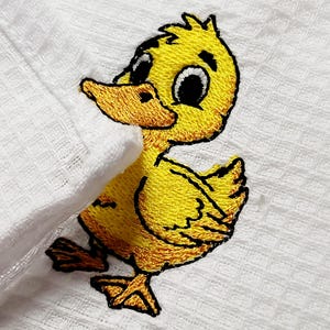 May include: Embroidered yellow duckling design on a white, textured fabric. The duckling has a detailed outline with black and orange accents, featuring large eyes and a friendly expression. The fabric appears to be a waffle weave.