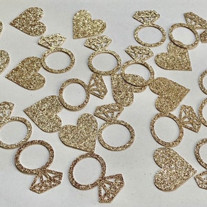 May include: Gold glitter confetti with heart and ring shapes. The rings have a diamond design.