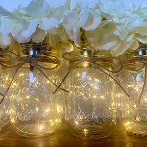 May include: Four clear glass mason jars with white string tied around the top and a white hydrangea flower arrangement on top. Each jar contains a string of white fairy lights.