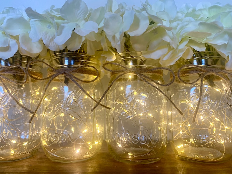 May include: Four clear glass mason jars with white string tied around the top and a white hydrangea flower arrangement on top. Each jar contains a string of white fairy lights.