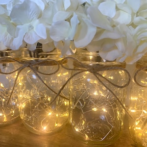 May include: A row of clear glass mason jars with white faux hydrangeas and string lights inside. The jars are tied with twine and the lights are lit, creating a warm glow.