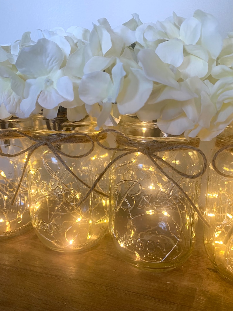 May include: A row of clear glass mason jars with white faux hydrangeas and string lights inside. The jars are tied with twine and the lights are lit, creating a warm glow.