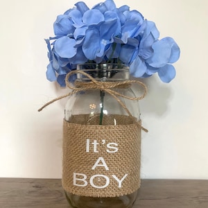 K&ouml;nnte beinhalten: Eine klare Glasvase mit einem Jutesband und einem Juteband, auf dem in wei&szlig;en Buchstaben "It's A Boy" steht. Die Vase ist mit hellblauen Blumen gef&uuml;llt.