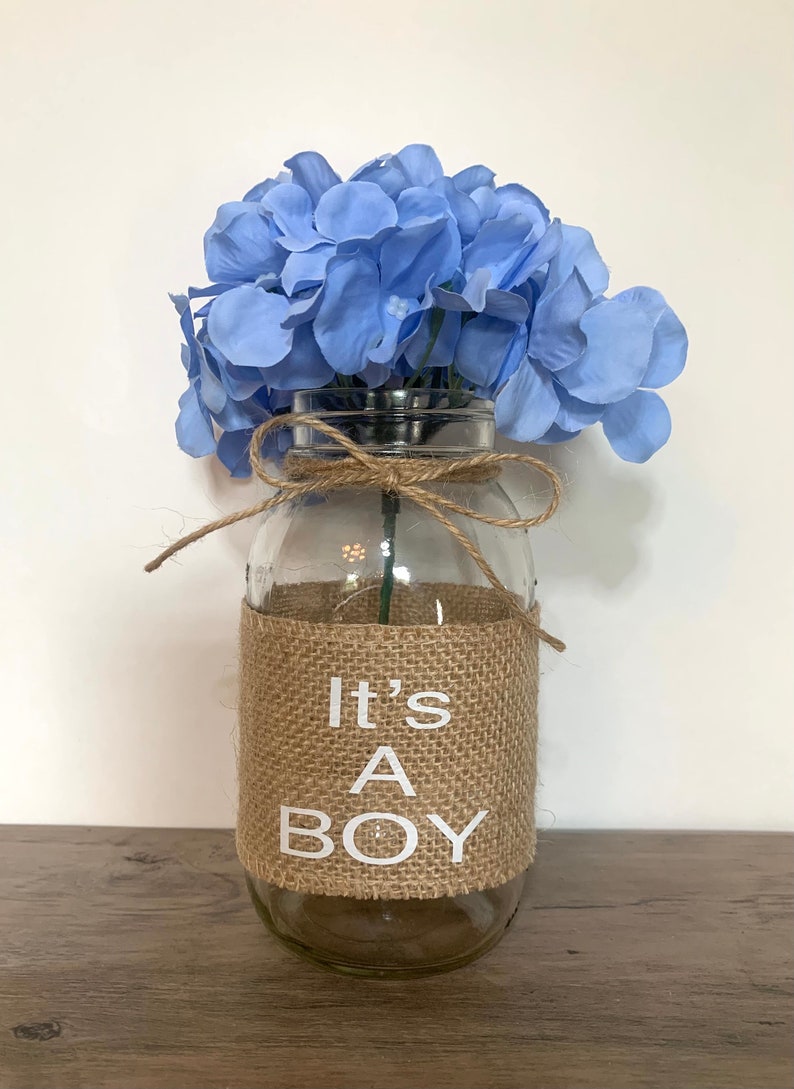 K&ouml;nnte beinhalten: Eine klare Glasvase mit einem Jutesband und einem Juteband, auf dem in wei&szlig;en Buchstaben "It's A Boy" steht. Die Vase ist mit hellblauen Blumen gef&uuml;llt.