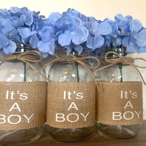 K&ouml;nnte beinhalten: Drei Glasvasen, die mit Sackleinen und dem Text "It's a Boy" in Wei&szlig; verziert sind. Jede Vase ist mit hellblauen Hortensien gef&uuml;llt.