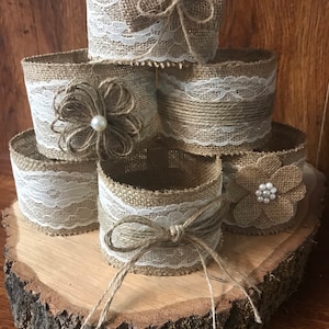 May include: A stack of six brown burlap and white lace cylinder vases with a bow and flower embellishments on each. The vases are arranged on a wooden slice.