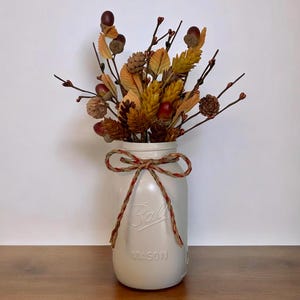 Peut inclure: Un bocal Mason en verre blanc cass&eacute; mat avec le texte en relief Ball et Mason, nou&eacute; avec un n&oelig;ud en ficelle d&eacute;corative. Le bocal est rempli d'un arrangement de feuillage d'automne artificiel, incluant des pommes de pin brunes et des glands rouges.
