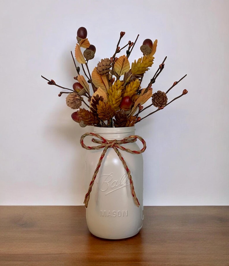 Peut inclure: Un bocal Mason en verre blanc cass&eacute; mat avec le texte en relief Ball et Mason, nou&eacute; avec un n&oelig;ud en ficelle d&eacute;corative. Le bocal est rempli d'un arrangement de feuillage d'automne artificiel, incluant des pommes de pin brunes et des glands rouges.