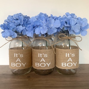 K&ouml;nnte beinhalten: Drei Glasvasen mit blauen Hortensienbl&uuml;ten und einem Juteband, das um die Oberseite gebunden ist. Jede Vase hat ein Juteband mit dem in Wei&szlig; gedruckten Text "It's A Boy".