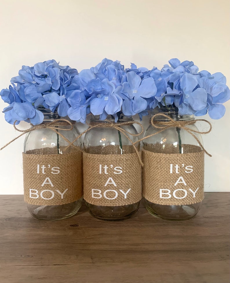 K&ouml;nnte beinhalten: Drei Glasvasen mit blauen Hortensienbl&uuml;ten und einem Juteband, das um die Oberseite gebunden ist. Jede Vase hat ein Juteband mit dem in Wei&szlig; gedruckten Text "It's A Boy".
