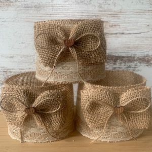 May include: Three brown burlap candle rings with white lace trim and a small wooden heart accent. The rings are stacked on top of each other.