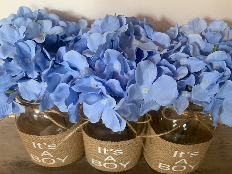 K&ouml;nnte beinhalten: Drei Glasvasen mit einem Jutestoffband und dem Text "It's a Boy" sind mit hellblauen Hortensien gef&uuml;llt.