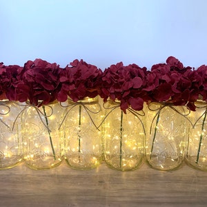 May include: A row of six clear glass mason jars with burgundy hydrangea flowers and string lights. The jars are sitting on a wooden surface.