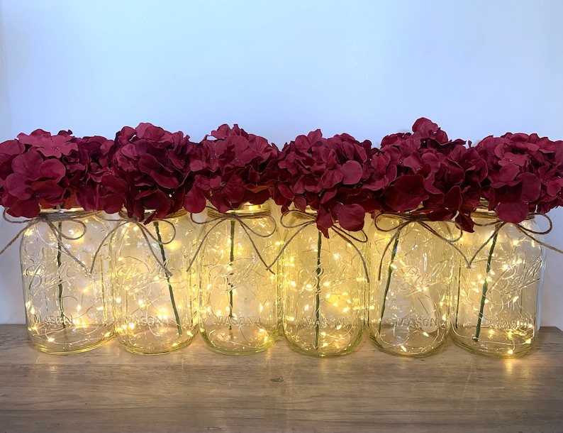 May include: A row of six clear glass mason jars with burgundy hydrangea flowers and string lights. The jars are sitting on a wooden surface.