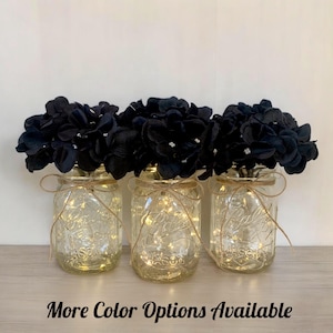 May include: Three mason jars with black silk flowers and fairy lights inside. The jars are tied with twine and the flowers are arranged in a bouquet.