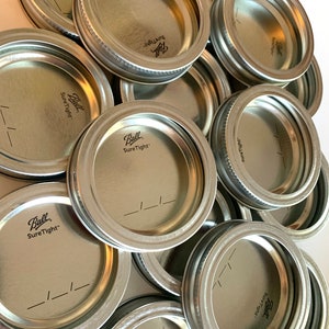 May include: A pile of silver metal lids for canning jars. The lids are labeled "Ball Sure Tight" and have a small groove around the edge.