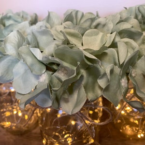 May include: A group of glass jars with string lights wrapped around them, decorated with light green artificial hydrangea flowers.