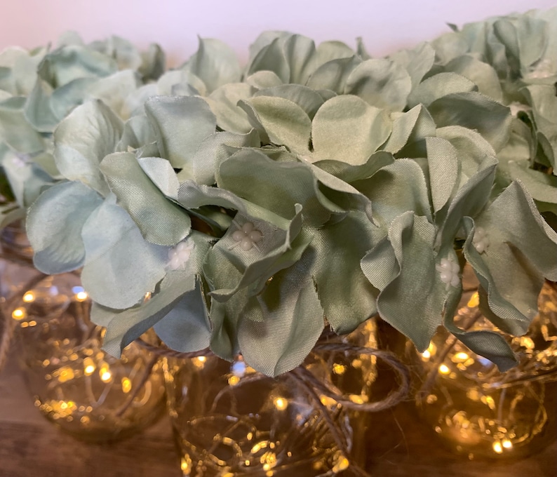 May include: A group of glass jars with string lights wrapped around them, decorated with light green artificial hydrangea flowers.