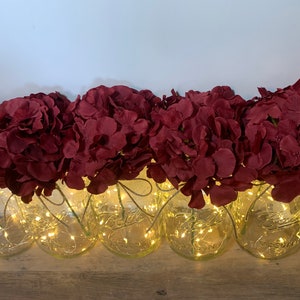 May include: A row of clear glass jars with a string of fairy lights wrapped around them. Each jar is filled with a bouquet of burgundy red artificial hydrangea flowers.