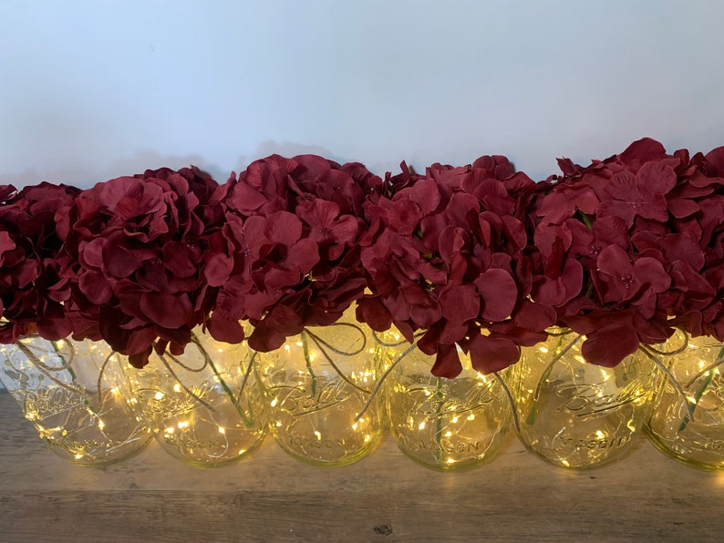 May include: A row of clear glass jars with a string of fairy lights wrapped around them. Each jar is filled with a bouquet of burgundy red artificial hydrangea flowers.